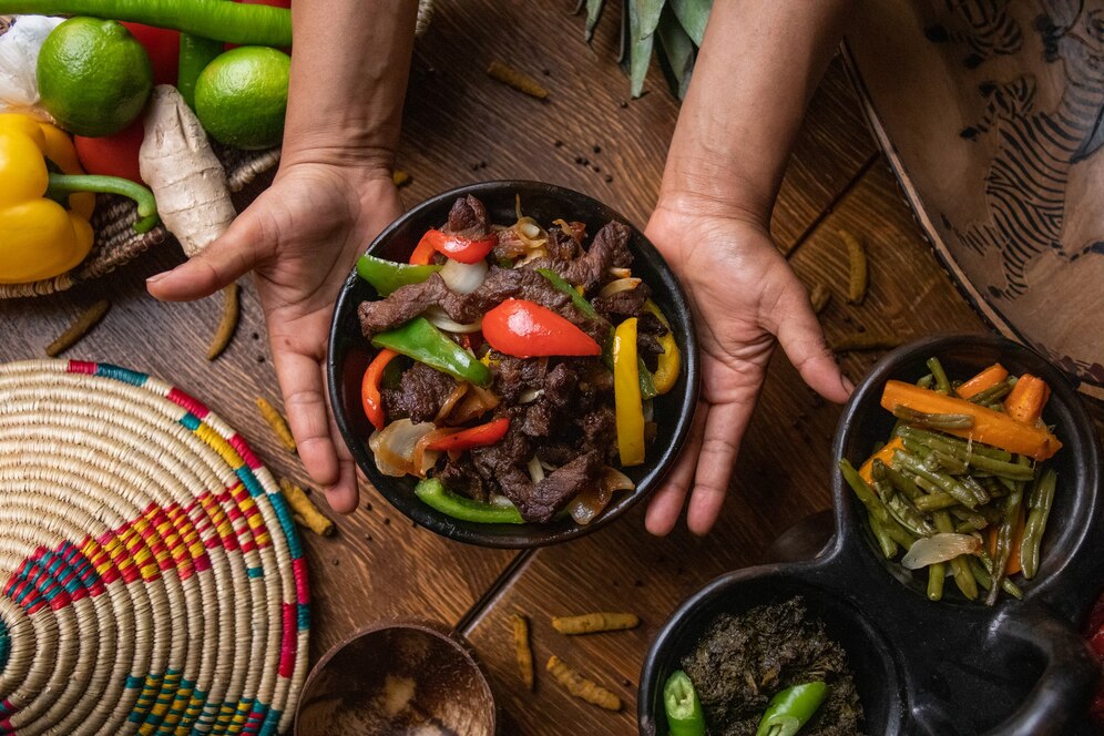 Hands serving stir-fried beef with bell peppers, onions, and vegetables on a rustic wooden table surrounded by fresh ingredients.