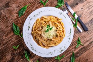 A plate of fettuccine pasta topped with grated cheese and herbs, served on a wooden table with cutlery and fresh basil leaves.