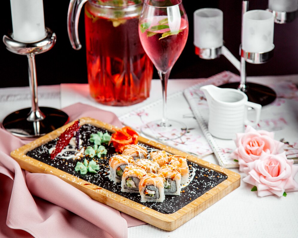 Elegant dining setup featuring sushi on a wooden board, pink roses, and drinks in stemmed glasses.