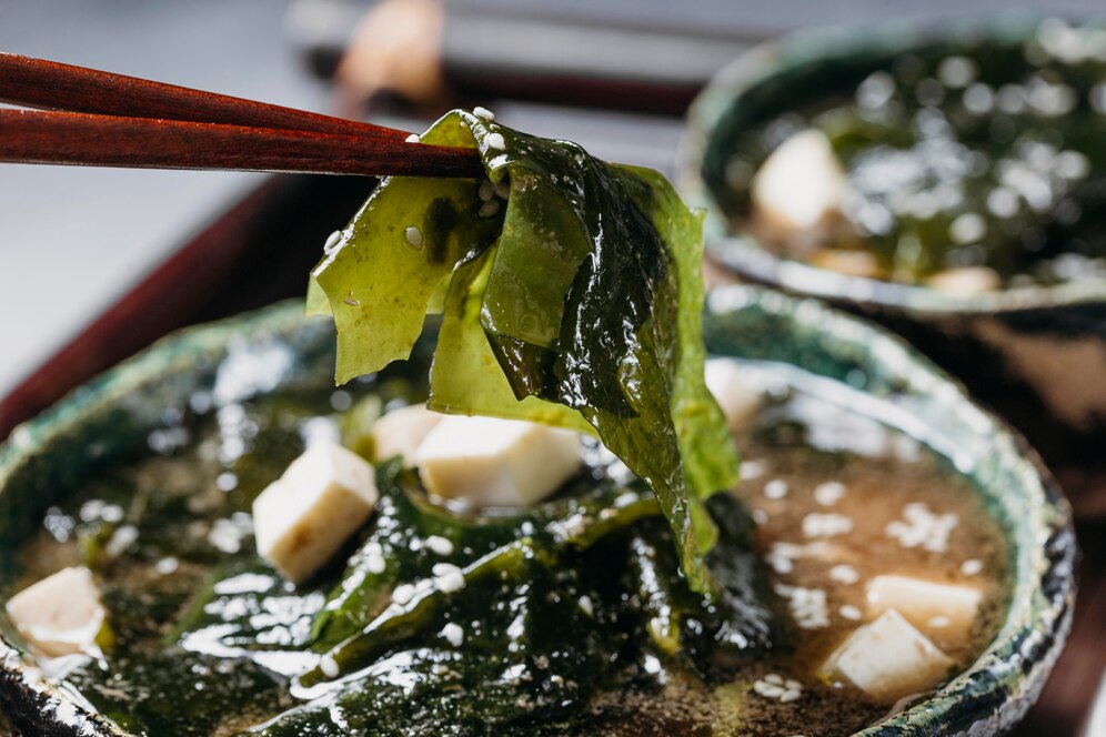 Chopsticks lifting seaweed from a bowl of miso soup with tofu and green seaweed.