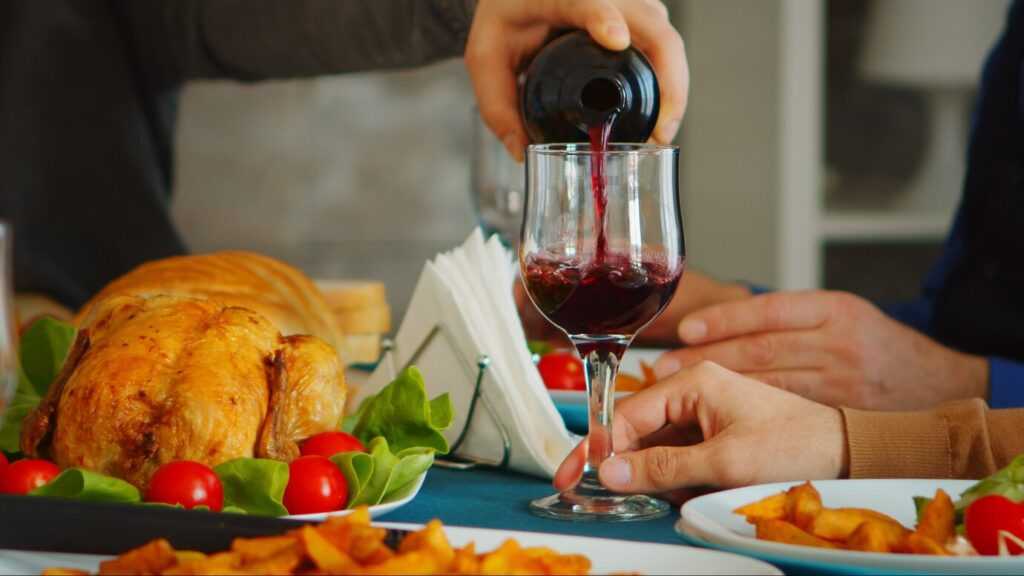 Someone pours red wine into a glass at a dining table with roasted chicken and vegetables.