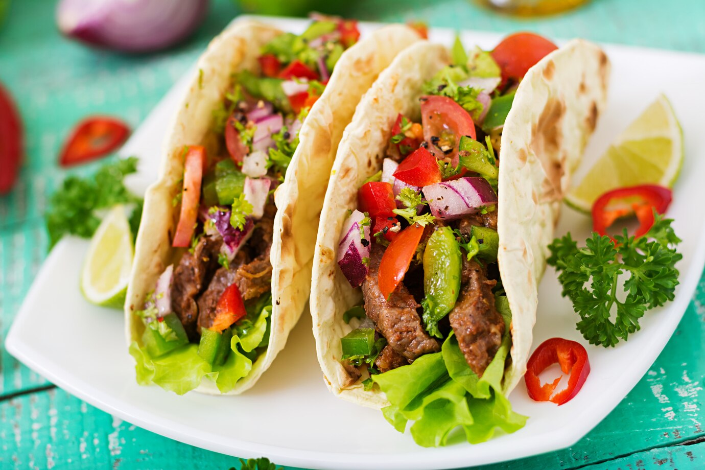 Three steak tacos filled with lettuce, vegetables, and vibrant toppings on a white plate, garnished with lime wedges.