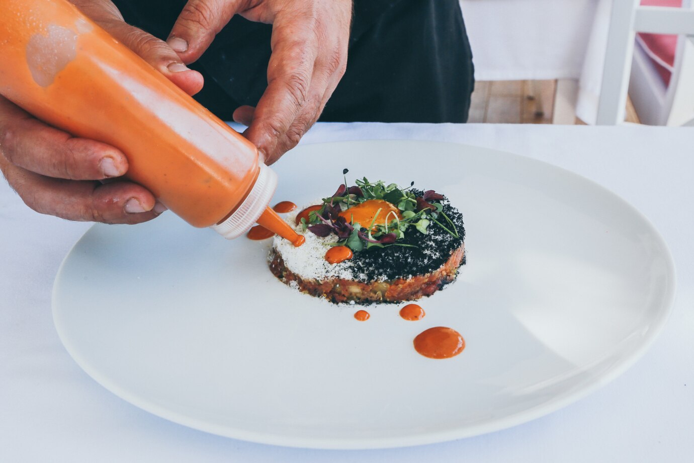 A chef drizzles orange sauce over a gourmet dish topped with microgreens and a vibrant egg yolk