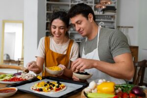 A man and woman in aprons prepare a pizza together in a home kitchen.