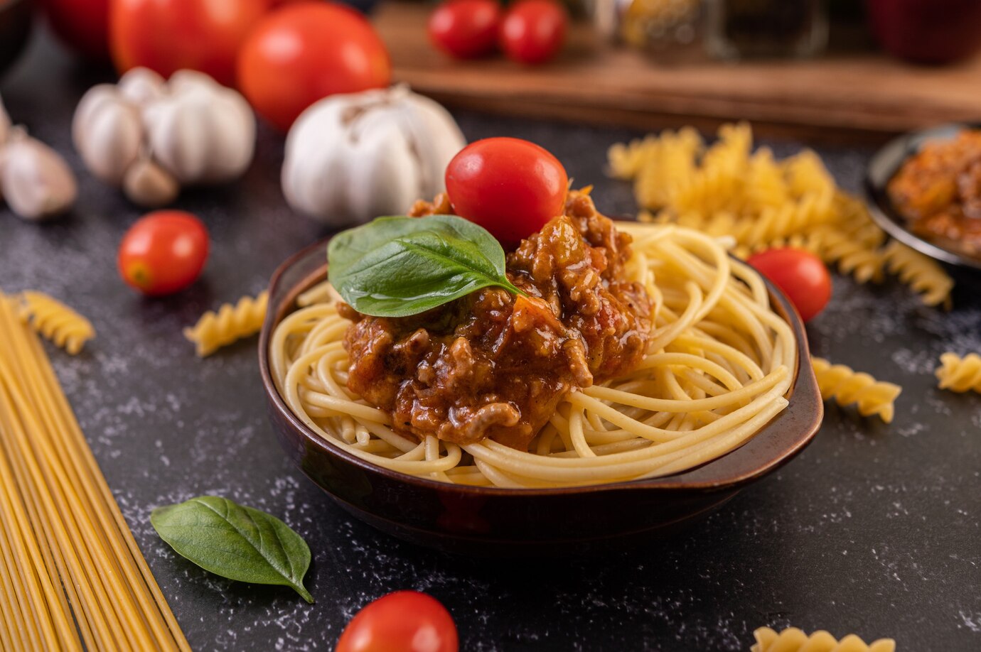 A bowl of spaghetti with meat sauce, garnished with a basil leaf and cherry tomato, surrounded by raw ingredients and pasta.