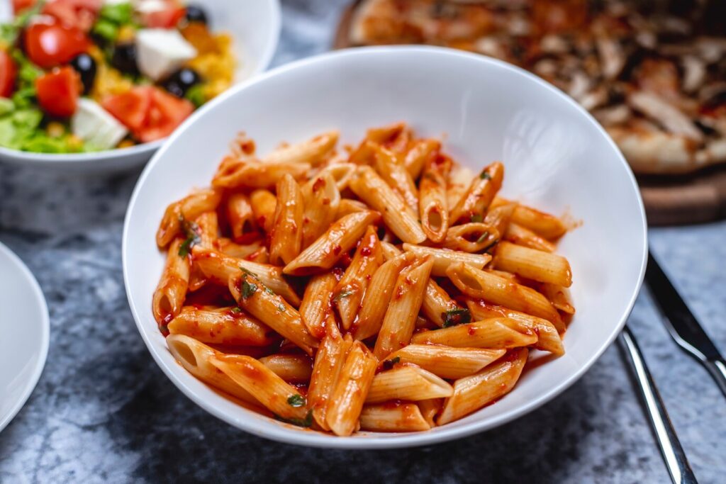 A bowl of penne pasta coated in a rich, red tomato sauce with herbs, served on a gray table.