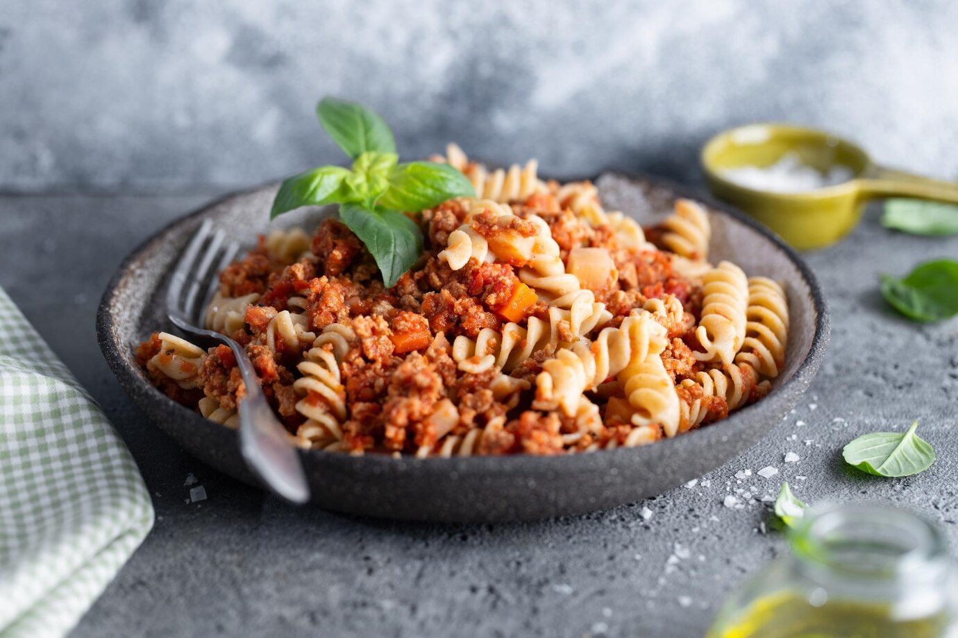 A bowl of fusilli pasta mixed with minced meat, tomato sauce, and topped with a basil leaf.