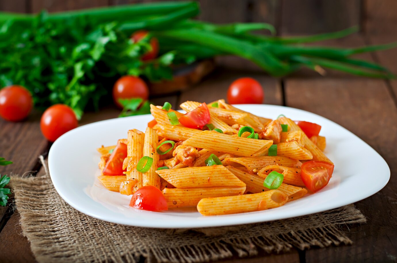 Plate of penne pasta with tomato sauce, cherry tomatoes, and green onions on a rustic wooden table.