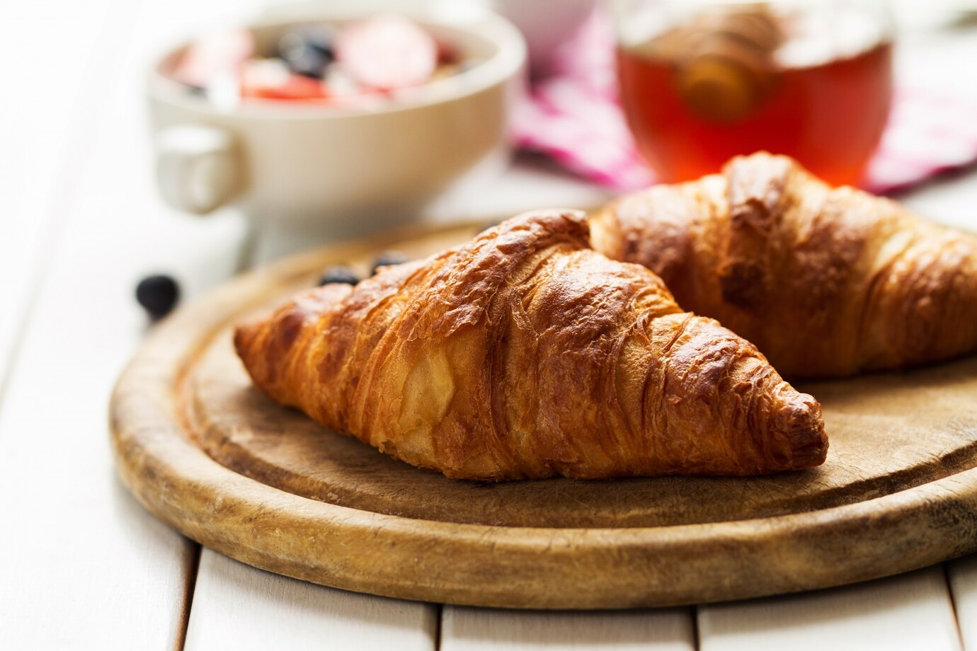 Two golden-brown croissants on a wooden board with a bowl of fruit and tea in the background.