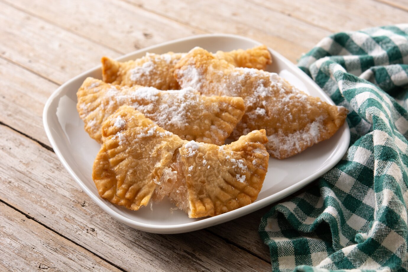 Plate of freshly fried pastries dusted with powdered sugar on a wooden table with a green checkered cloth.