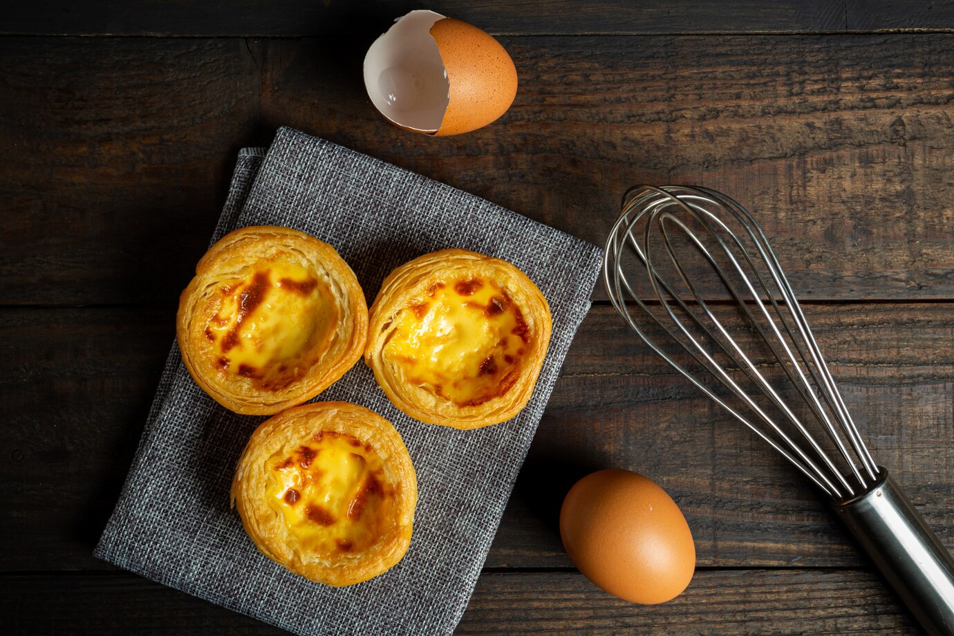 Three egg tarts with a whisk and eggs on a wooden table.