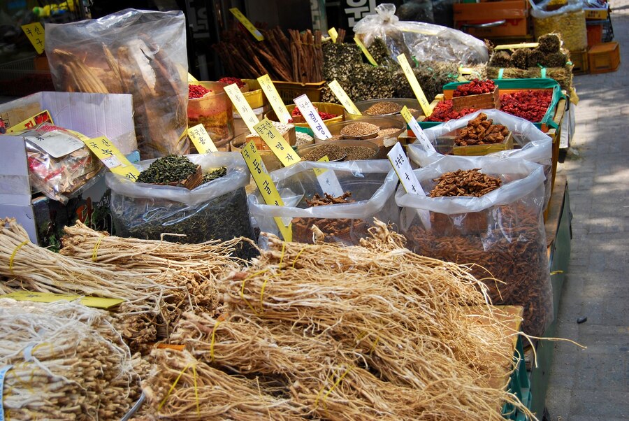 Variety of dried herbs, spices, and roots displayed in open containers at an outdoor market, with labels in a non-Latin script.