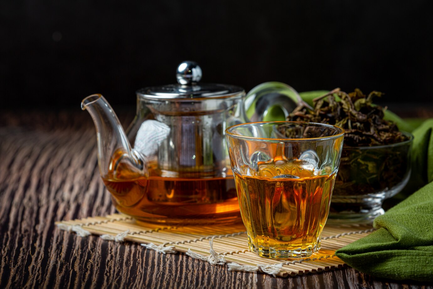 A glass cup of amber tea beside a glass teapot and a bowl of loose tea leaves