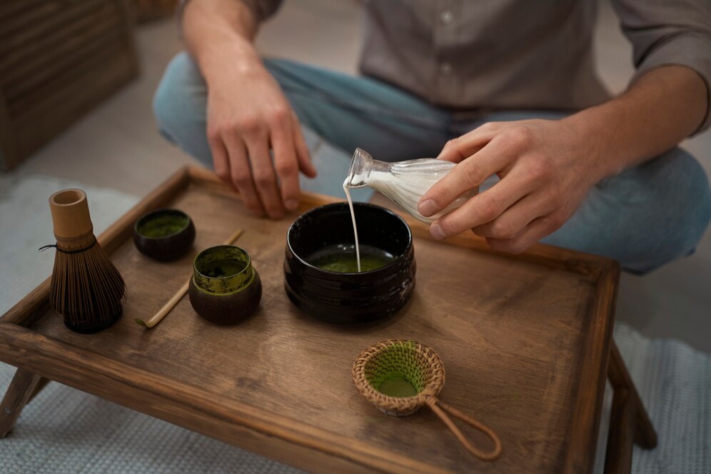 Person preparing matcha tea with a whisk and bowl on a wooden tray.