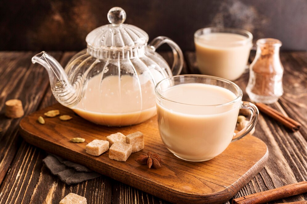 Glass teapot and cup of milk tea, sugar cubes, and spices on a wooden tray.